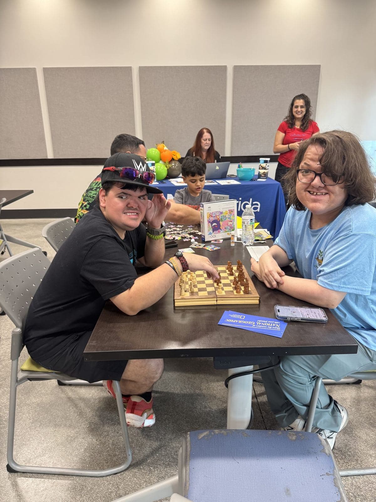 Two young ACES participants playing chess together at a community table while staff support others nearby